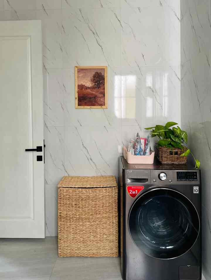 A clean, modern laundry space featuring a white wall and door background with a washing machine, rattan storage basket, and a plant.