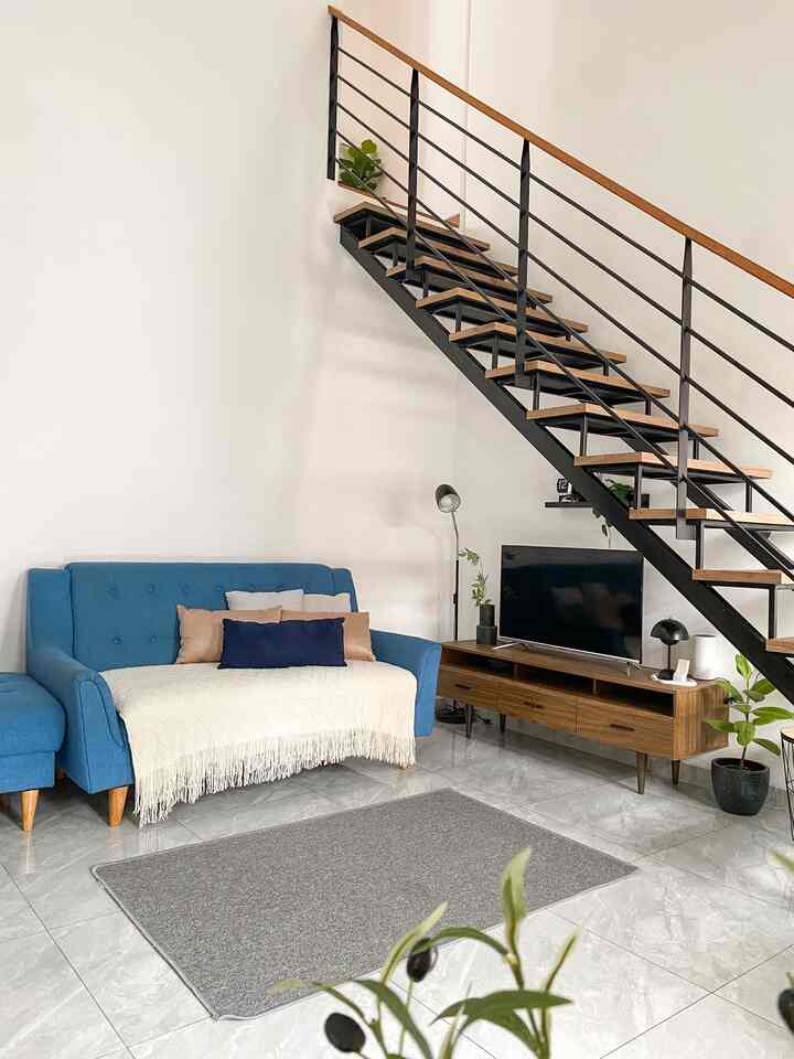 Modern living room with gray tiled floor, wood-toned TV stand, blue sofa with white throw blanket, and staircase