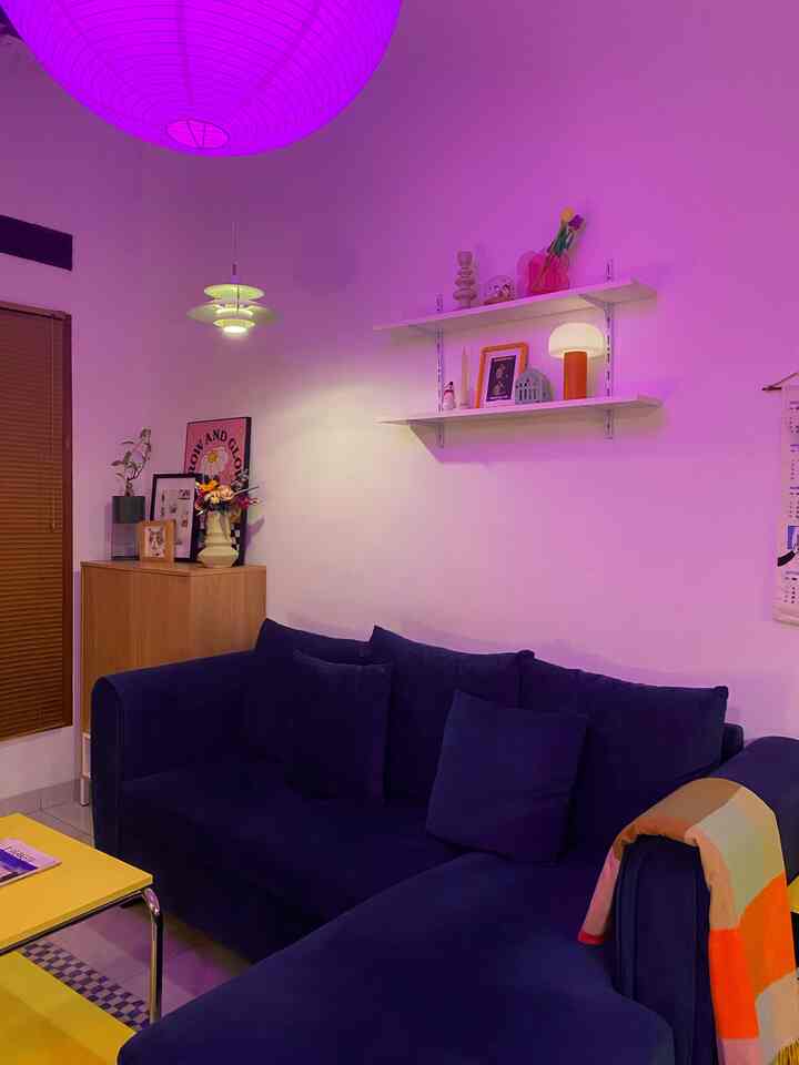 Living room with white walls softly lit by purple lighting, featuring a blue sofa and wooden shoe cabinet creating cozy ambiance