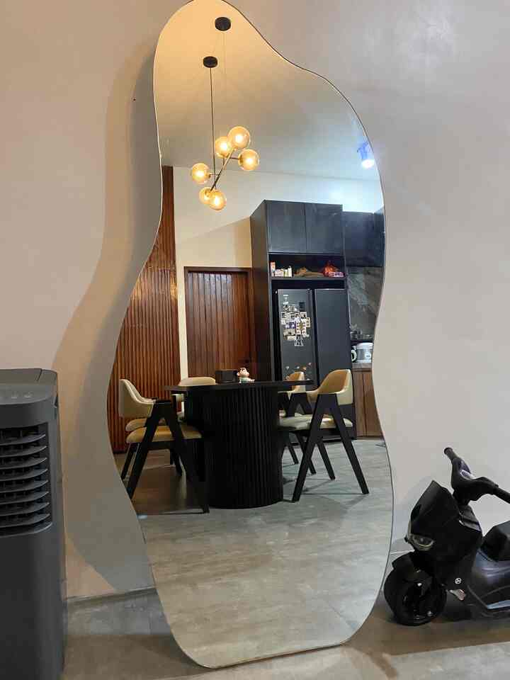 A uniquely shaped large floor mirror reflects a dining room with a curved black dining table and beige chairs adjacent to a kitchen area.