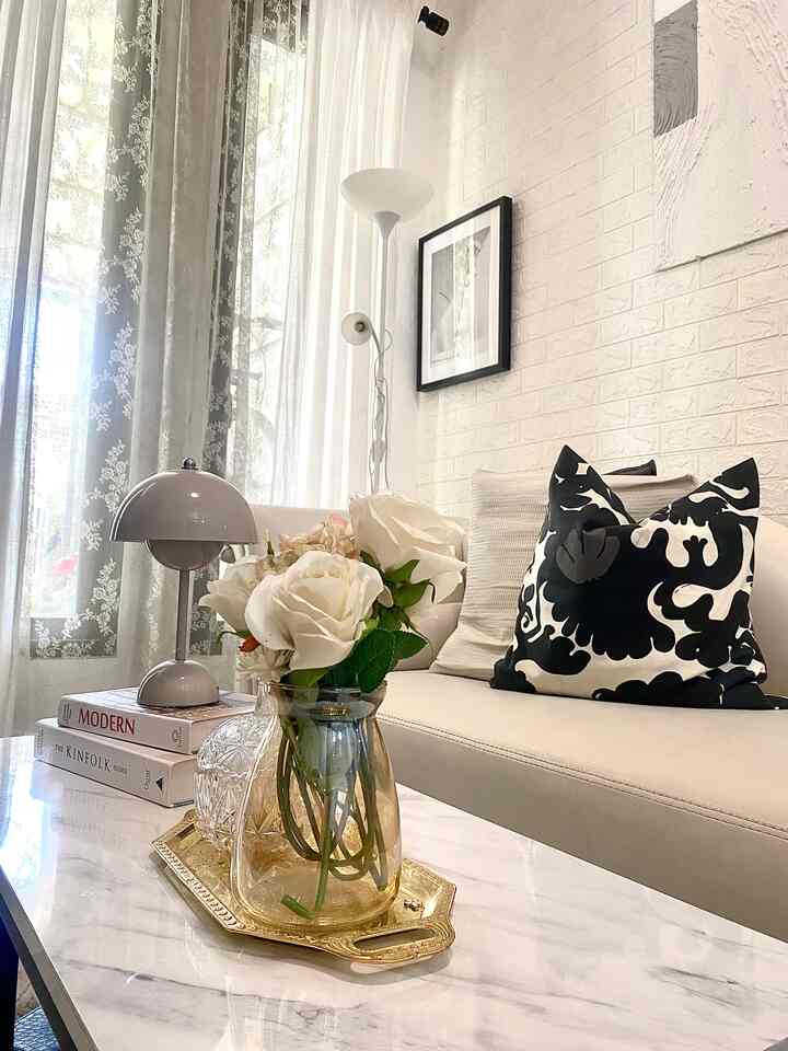 A white and beige toned living room featuring a beige sofa with black patterned cushions, a table with a gold tray centerpiece, and a floor lamp for cozy ambiance