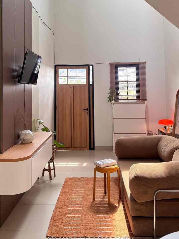 A warm wood-toned and beige living room featuring a large sofa bed, orange carpet, small stool, and a wall mirror with a simple style