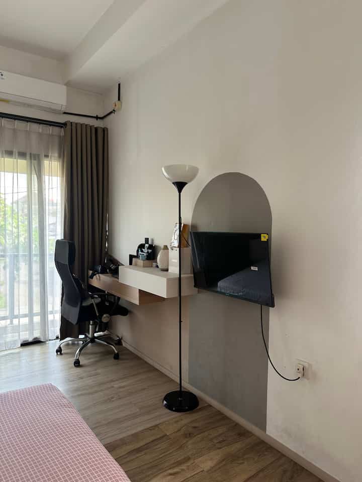 Small modern bedroom with white walls and brown wooden floor, featuring a home office area, floor lamp, and minimal furnishings
