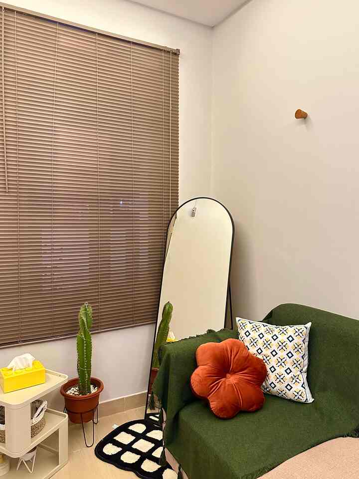 Living room with white walls and brown blinds featuring a green sofa, orange cushions, and a full-length mirror creating a cozy atmosphere