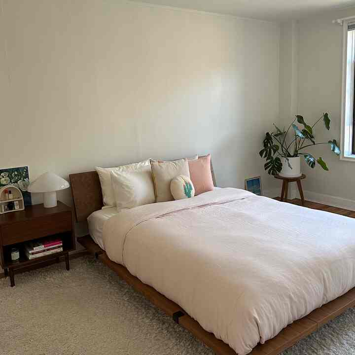 Soft pink bedding and natural wood furniture in a white-toned bedroom with a green plant adding natural touch