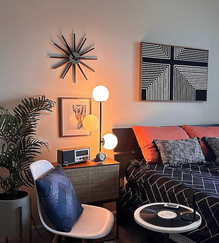 A mid-century modern bedroom with white walls and black bed, accented by orange pillows and a glowing floor lamp creating a cozy atmosphere