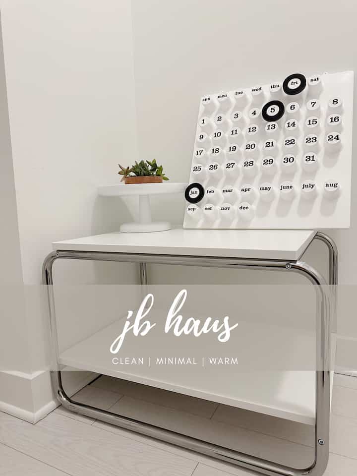 Minimalist living room corner in white and silver tones featuring a console table, perpetual calendar, and small plant with a clean atmosphere