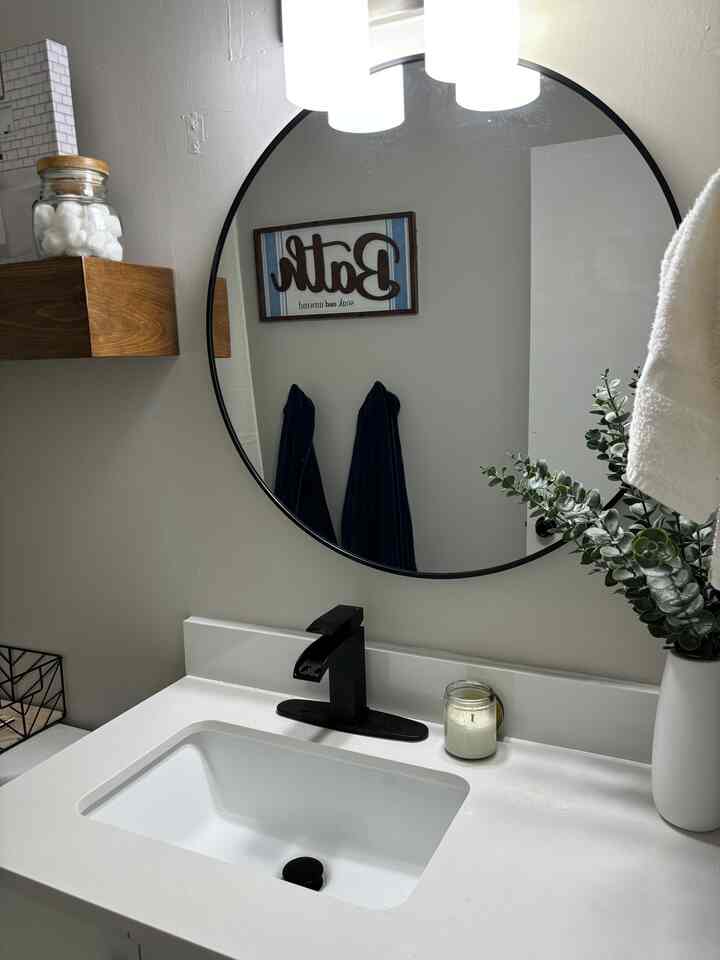 A white and black toned bathroom featuring a round mirror, sink, wooden shelf, and green plant creating a clean look