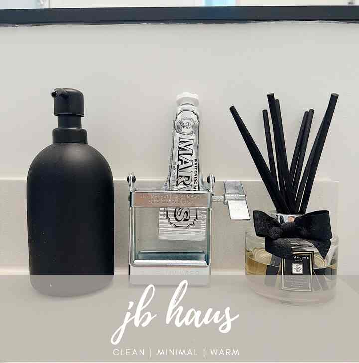 Minimal bathroom space with white background featuring a black soap dispenser on left, a metal tube wringer center, and black stick diffuser right