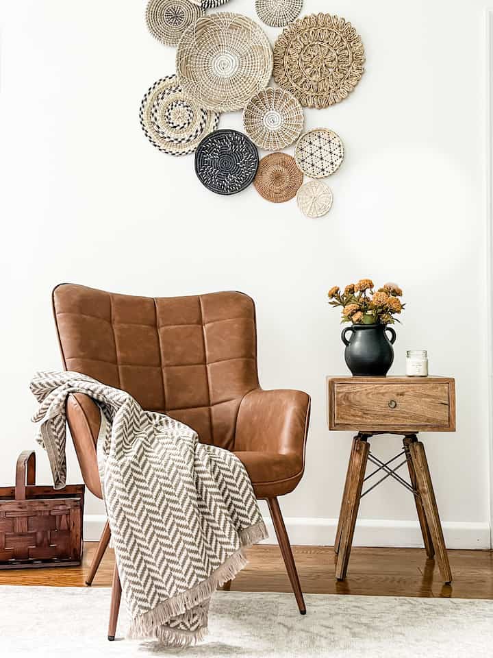 Brown armchair with patterned throw blanket and wooden side table featuring a vase with flowers and candle against white wall in a living room