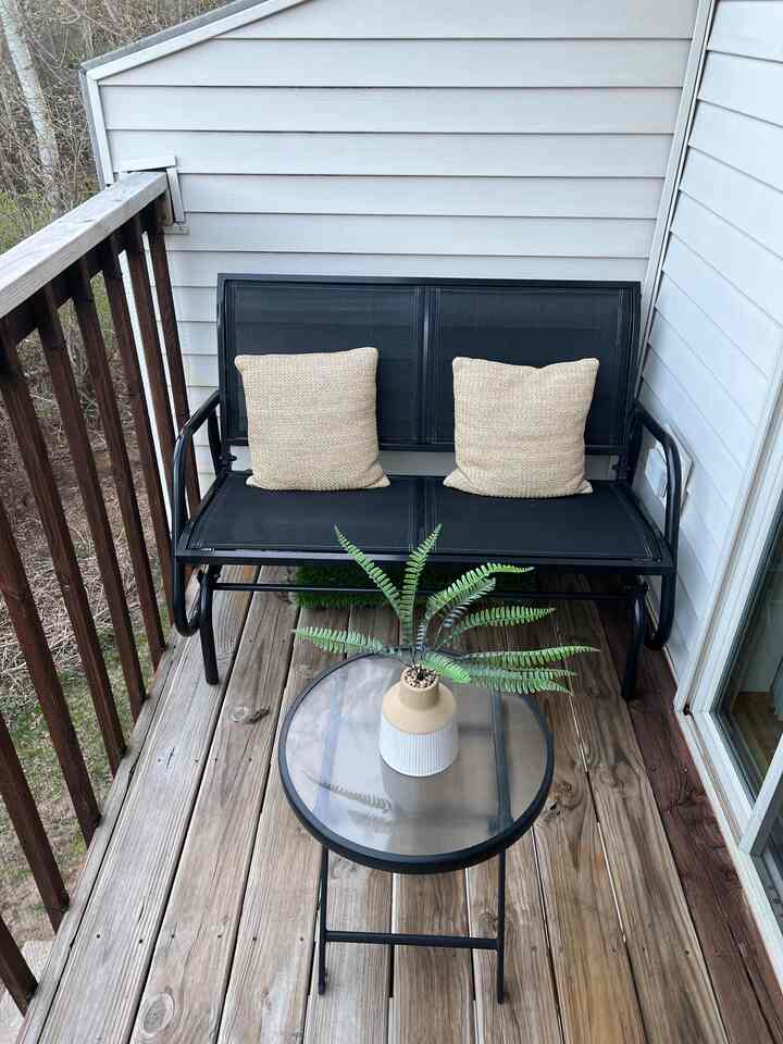 Black framed outdoor glider chair with beige cushions, glass top side table on wood tone apartment deck