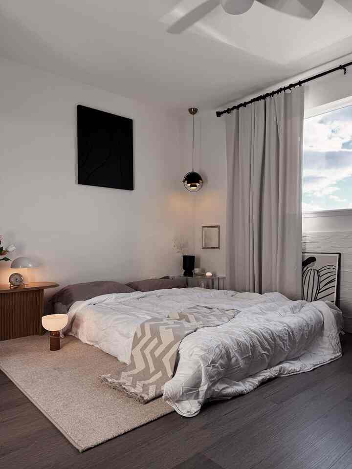 A white and beige toned bedroom featuring a large cozy bed with pillows, table clock, and hanging pendant light creating a warm atmosphere