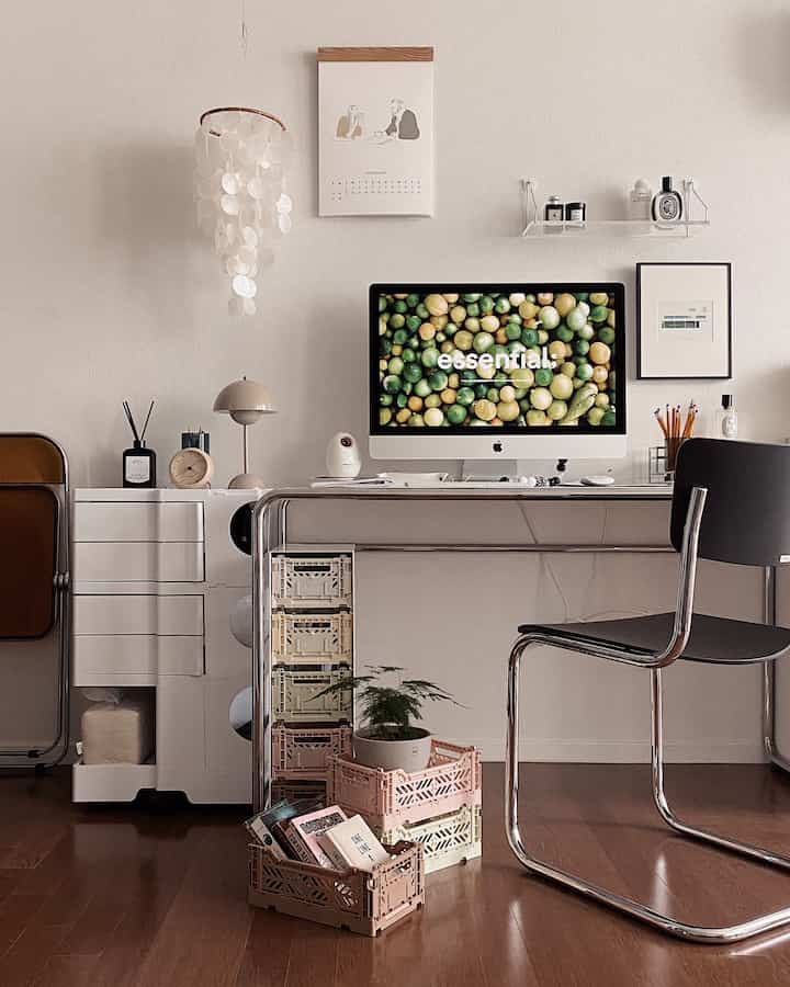 Modern home office with white walls and wood tone flooring, featuring a metal desk with monitor centered and organized storage boxes on the left