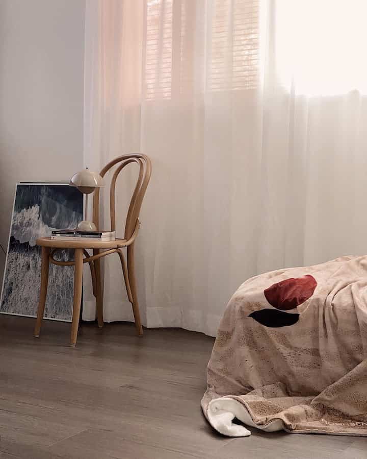 Natural toned wooden floor with Ton chair, cream table lamp, beige blanket beside bright curtains in a living room corner
