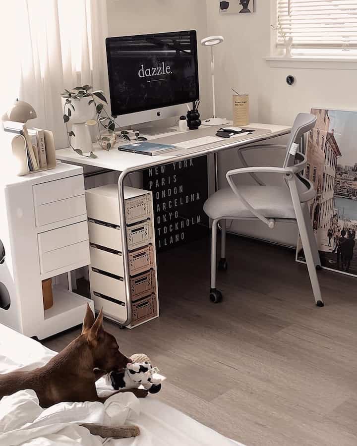 Modern home office with white and wood tones, featuring iMac on desk and gray office chair, brown dog playing with toy on floor