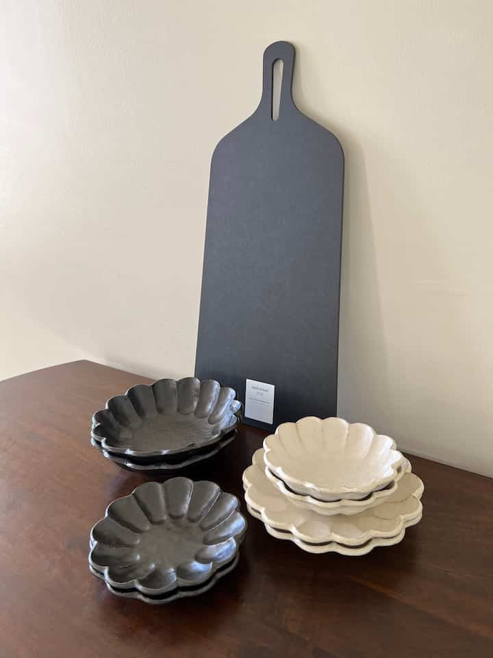 Natural wood tone dining table featuring dark gray cutting board and stacked black and white flower-shaped bowls and plates in a tidy table setting