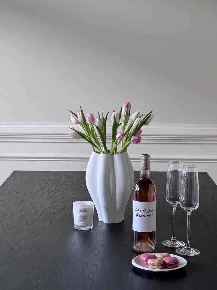 A simple modern dining room with white walls and a black dining table featuring pink tulip vase, wine bottle, and candle setup