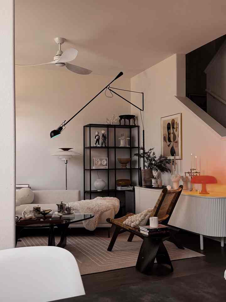 Bright white and brown toned Mid-Century Modern living room featuring sofa, rattan armchair, black shelving unit, and layered lighting