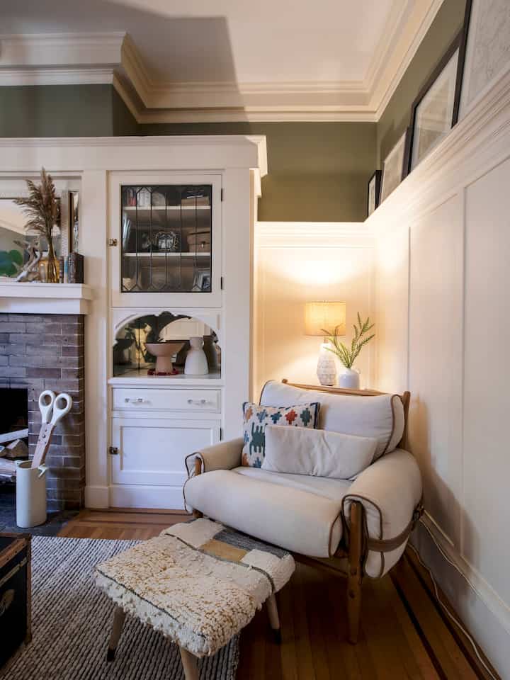 Cozy living room corner with natural brown wood floor, white paneled walls, and a cream armchair featuring a patterned cushion