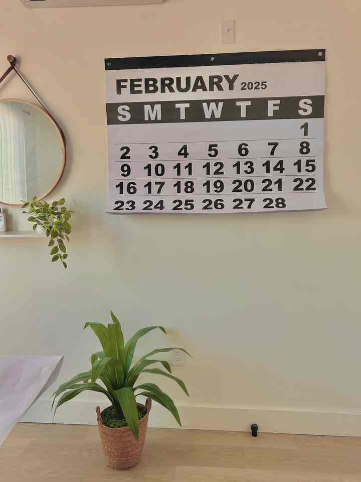 Bright white-toned home office space featuring a large wall calendar, round mirror on the left, and a brown basket plant on the wooden floor creating a clean interior