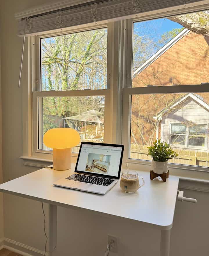 White-toned home office space featuring a desk by large windows, laptop, table lamp, and plant creating a warm work setting
