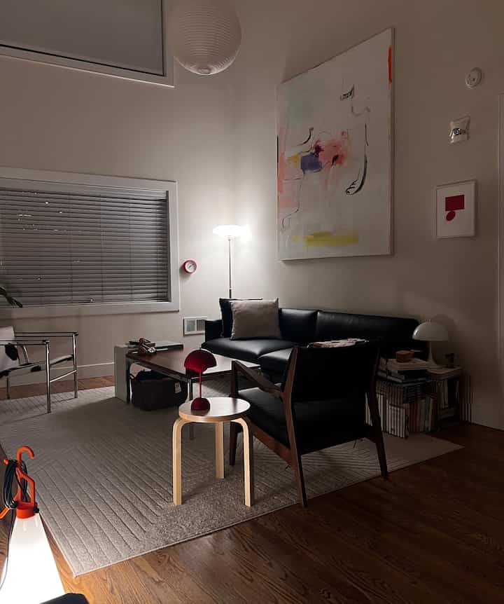 A white-walled living room with wood-toned flooring featuring black sofa and armchair, light gray rug, and red accent lamp in a Nordic modern style