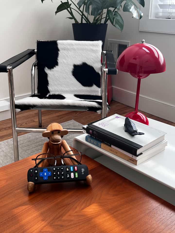A modern living room with white walls and brown wooden coffee table, featuring Cassina armchair, red table lamp, and small monkey objet decor