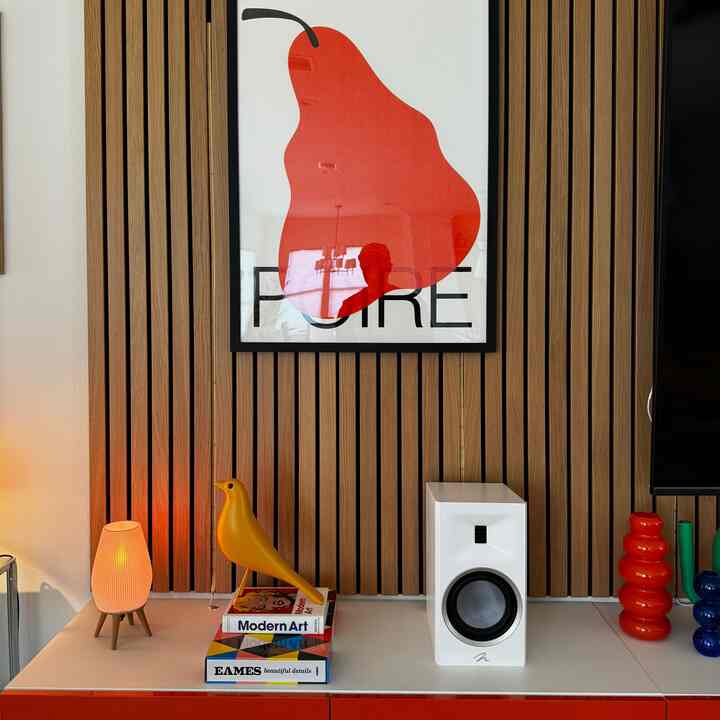 Modern living room corner with bright wood panel wall featuring large red pear artwork, white shelf displaying yellow bird object, white speaker, and red vases