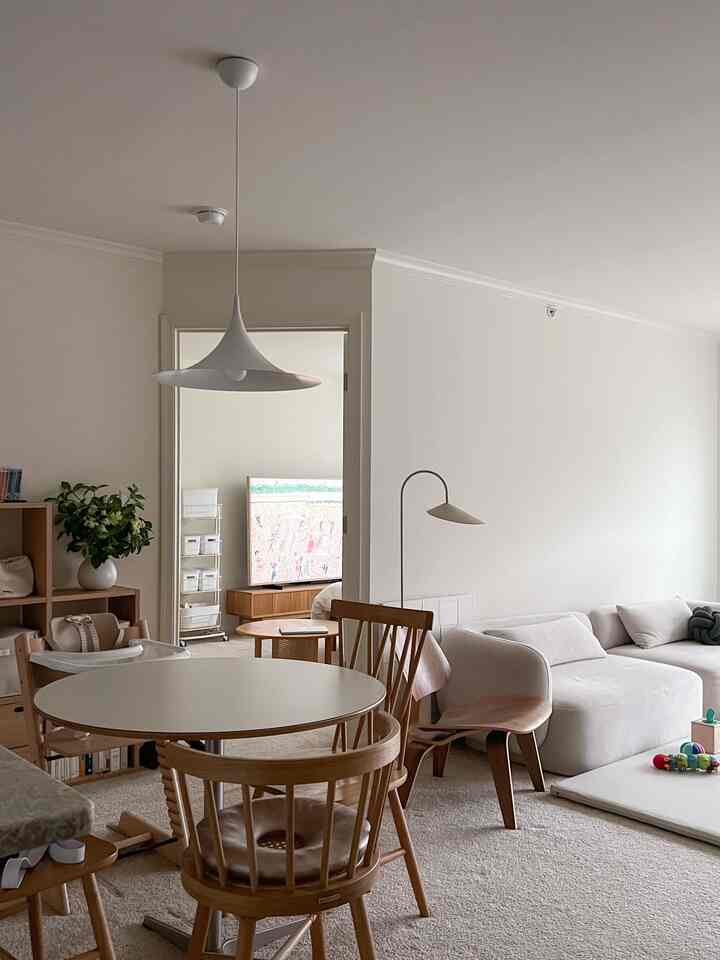 A cozy living room in soft beige and natural wood tones featuring a round dining table, large sofa, and children's play mat