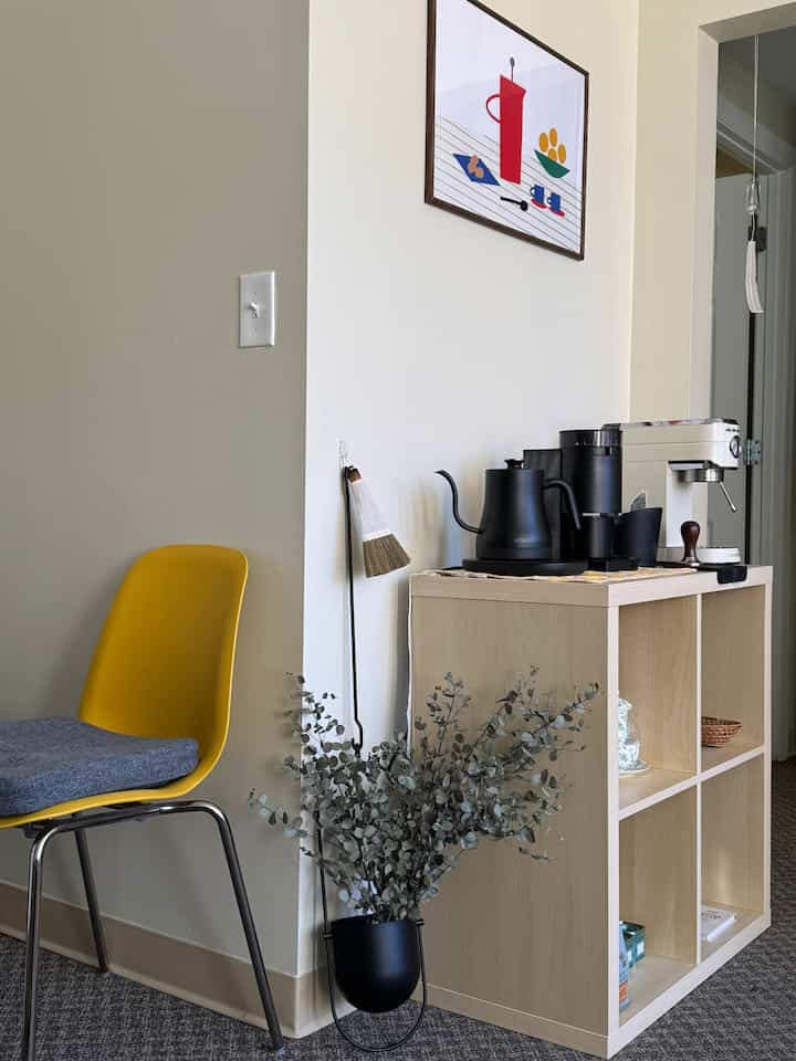 Natural toned home cafe corner with a yellow chair and wooden shelf featuring coffee equipment, creating a simple and cozy atmosphere