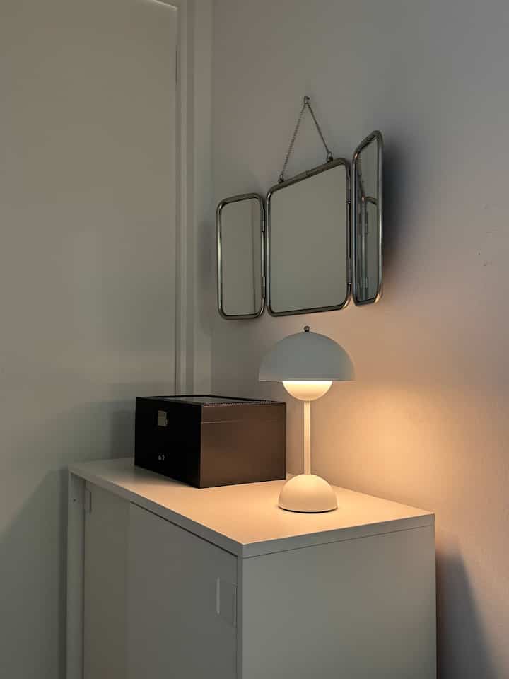 White and black toned cozy corner featuring a simple shoe cabinet, vintage tri-fold mirror, and soft table lamp lighting