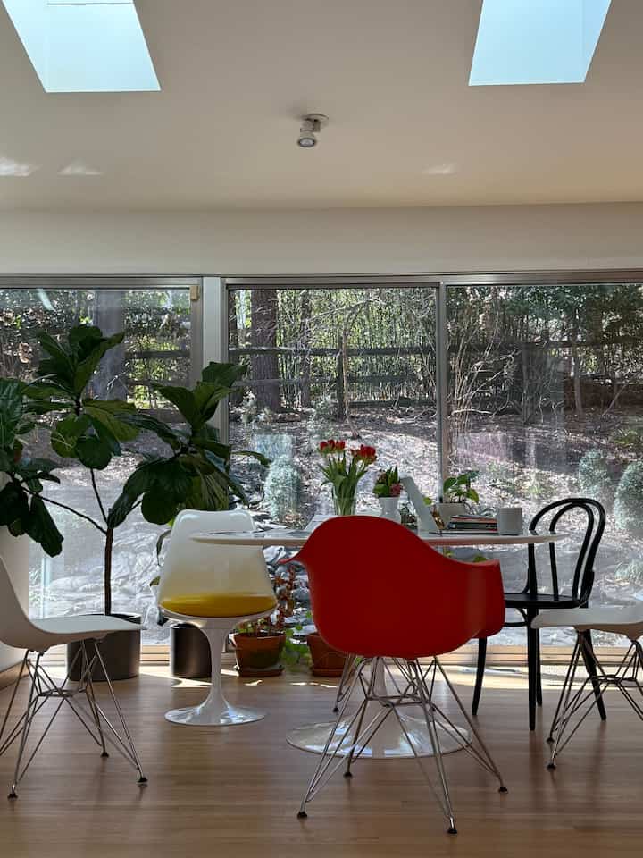 Bright dining room with white and natural wood tones, featuring a red armchair and diverse chairs, illuminated by ample natural light