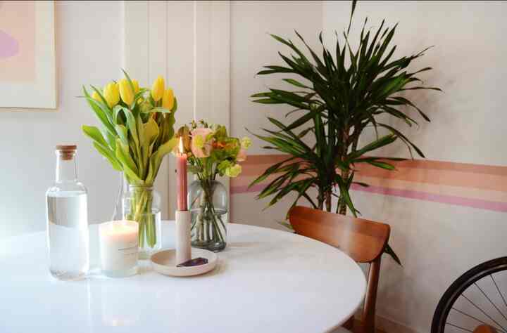 White-toned dining room featuring a white round dining table, wooden dining chair, yellow tulips in vases, and candles creating a natural atmosphere