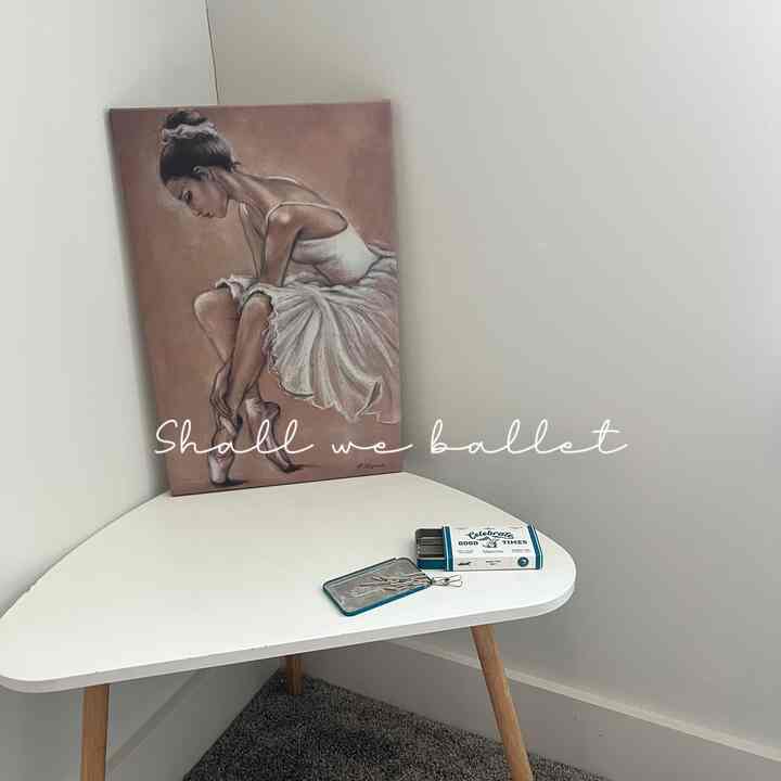 A cozy small corner room with white walls, natural wood-tone legged corner table and beige ballerina painting