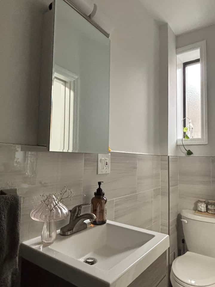 A cozy bathroom in soft beige and white tones featuring a transparent mushroom-shaped vase and brown dispenser on the sink