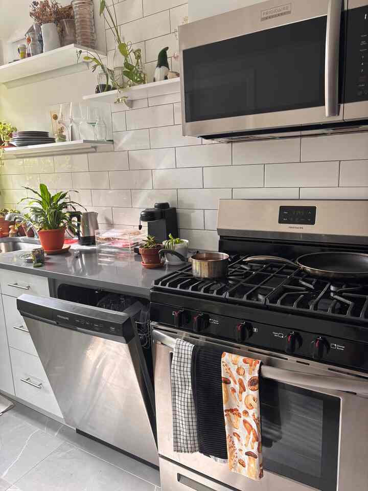 Modern apartment kitchen with white tile backsplash and stainless steel appliances, featuring plants on the sink counter and towels hanging on the oven