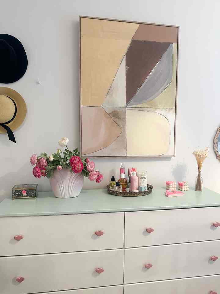 White and natural tone bedroom with a dresser featuring pink vase and abstract painting, creating a cozy atmosphere