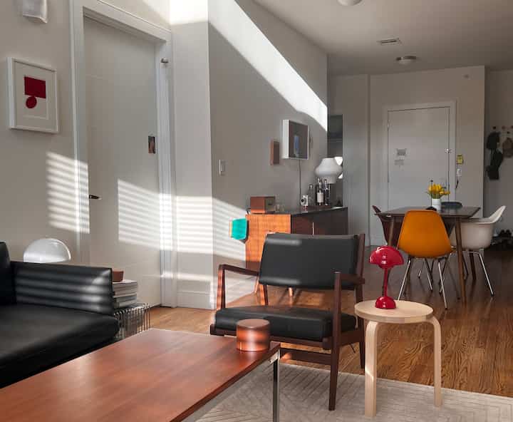 Modern living room with white walls and wood tone floor, featuring black sofa and armchair, wooden stool with red table lamp