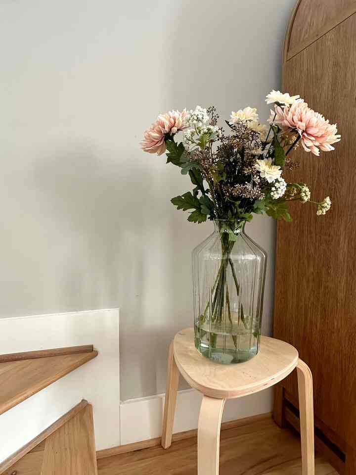 Natural wood tone corner space featuring a stool topped with a clear vase holding soft pink and white faux flowers, presenting a simple interior