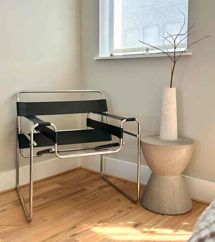 Bright natural corner workspace featuring a silver-frame black leather armchair and concrete side table near window with blinds