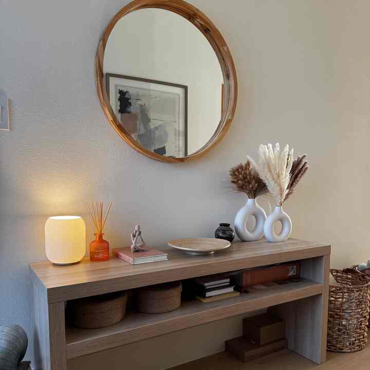 Wood tone and beige entrance featuring a round wood mirror and console table with table lamp and vases, creating a cozy atmosphere