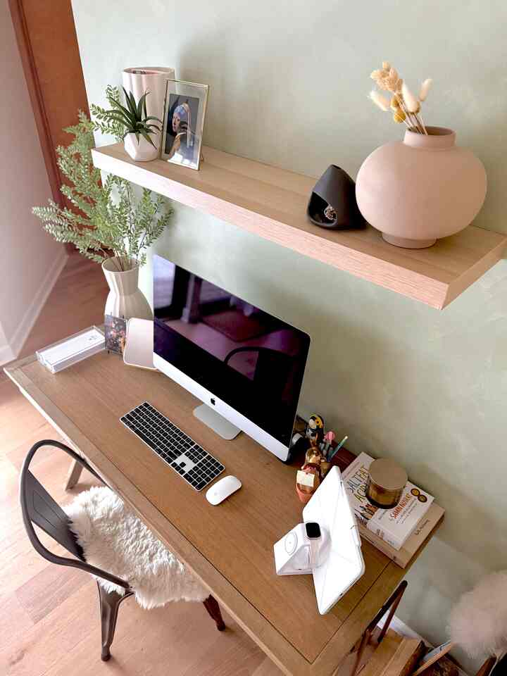 Cozy home office with a muted green wall and natural wood-toned desk featuring an iMac and decorative shelf