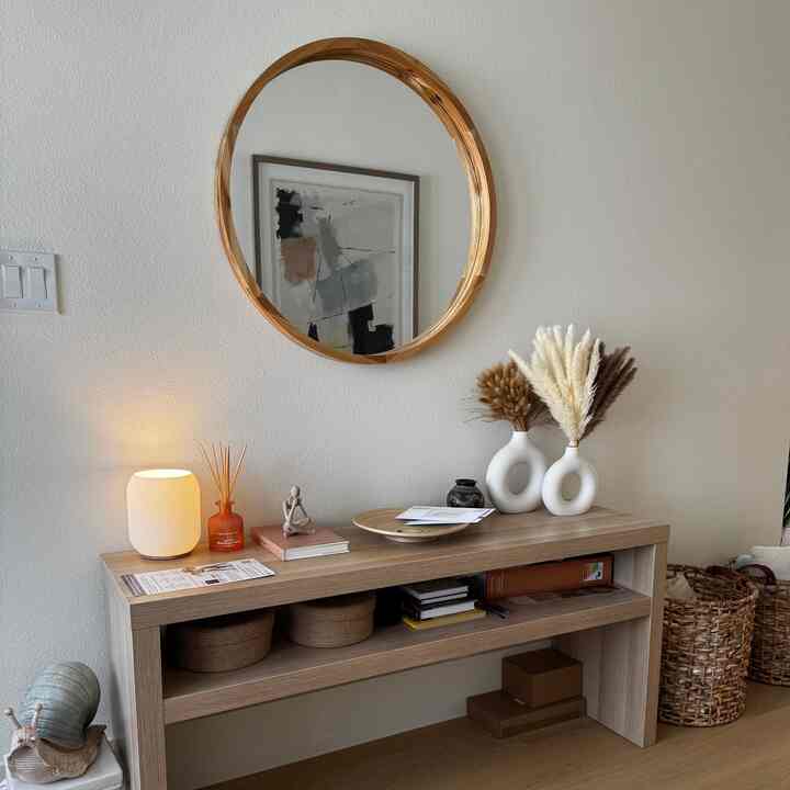 Soft oak-toned entrance featuring round wood mirror, table lamp, and white vases creating a cozy atmosphere
