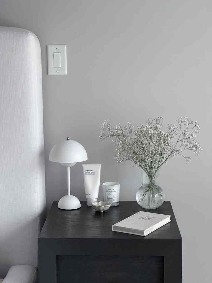Minimalist bedroom nightstand in white and gray tones featuring a table lamp, vase with flowers, candle, and a book creating a calm atmosphere