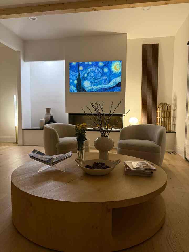 This #livingroom features a #modern and #minimalist design with warm, neutral tones. The space is illuminated by soft ambient lighting, creating a cozy atmosphere. A large, circular wooden coffee table with built-in storage serves as the focal point, complemented by two curved, cream-colored armchairs that add elegance and comfort.

A digital art display on the wall showcases Van Gogh’s “Starry Night”, adding a vibrant pop of color and an artistic touch. The fireplace, framed with a sleek, modern design, contributes to the room’s warmth. Decorative elements like a ceramic vase with yellow flowers, a white sculptural vase with branches, and curated coffee table books add layers of visual interest.

The #woodflooring and exposed ceiling beam enhance the natural and earthy feel of the space. A tall, vertical wood panel on the right introduces texture, while the recessed lighting and strategically placed floor lamp provide balanced illumination. The overall aesthetic is a perfect blend of contemporary sophistication and inviting comfort.
