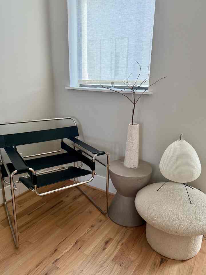 Simple mid-century modern living room corner with bright wood-tone flooring and gray walls, featuring a black armchair, light gray side table, and white stool