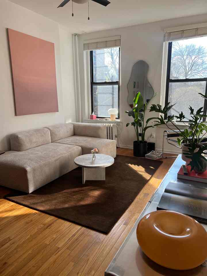 Bright living room with soft beige sectional sofa and brown rug, featuring large windows, natural light, and leafy plants in a natural modern style