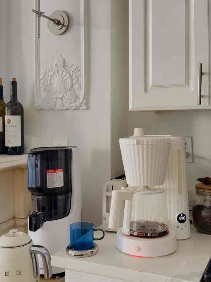 White-toned kitchen space featuring a Plissé drip coffee maker and Smeg electric kettle forming a minimalistic home cafe setup