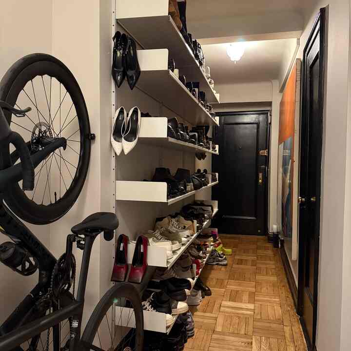 A narrow entrance hallway with white wall-mounted shelves filled with shoes, brown wooden parquet floor, and a bicycle positioned on the left side