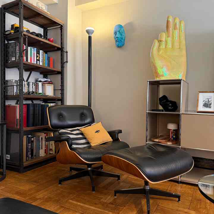A beige-toned living room with brown wood flooring featuring a Mid-Century Modern Eames Lounge Chair and Ottoman, bookshelf, and floor lamp creating a cozy reading space